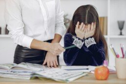 Upset little girl sitting near crop woman in classroom